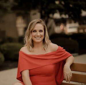 Chelsea Ohlemiller, smiling, seated outdoors on a bench in a red off-the-shoulder dress
