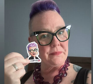 Tracey Piparo, with purple hair and glasses, holding a small cartoon avatar pin of herself