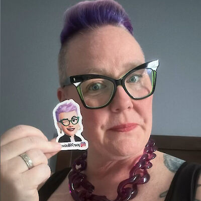 Tracey Piparo, with purple hair and glasses, holding a small cartoon avatar pin of herself