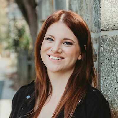 Headshot of Sarah Adams, smiling with long auburn hair, against a stone wall
