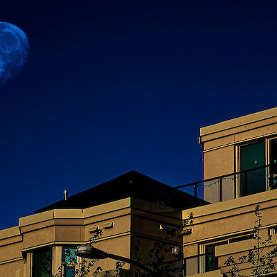 An almost-full moon glowing in a deep blue sky above a modern apartment building at dusk