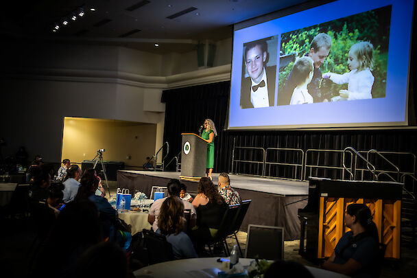 A speaker at a podium on a darkened stage, with photos of a young donor projected on a large screen behind her and an audience seated at tables