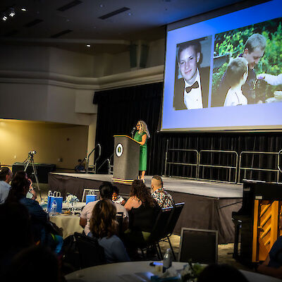 A speaker at a podium on a darkened stage, with photos of a young donor projected on a large screen behind her and an audience seated at tables
