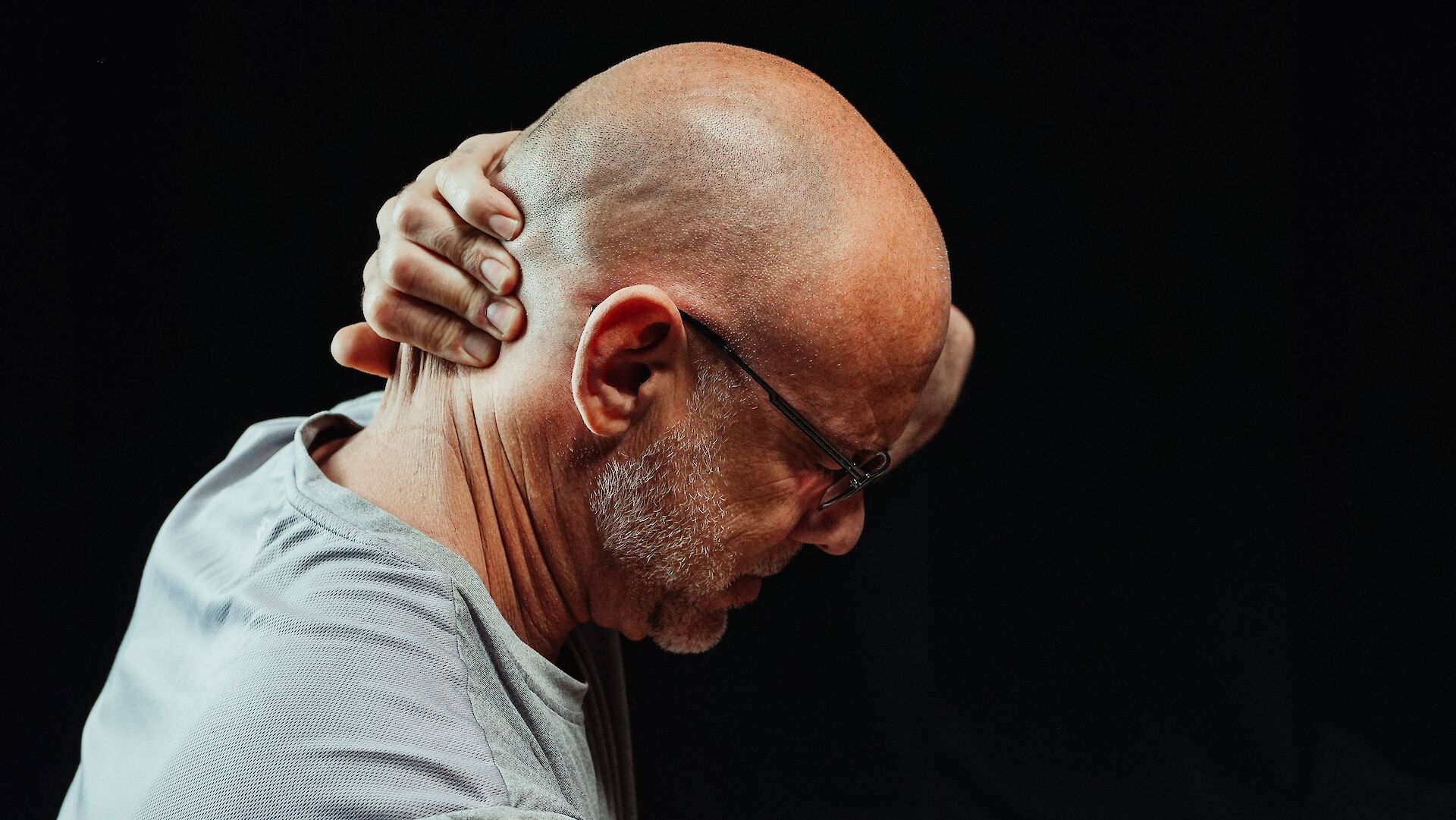 A side profile of a man looking downward while gripping the back of his neck in tension, representing the physical toll and bodily strain that comes from suppressing emotions during grief.