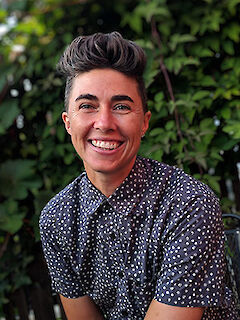 Katrina Spade, smiling, with short dark hair styled up, wearing a polka-dot shirt, in front of green foliage