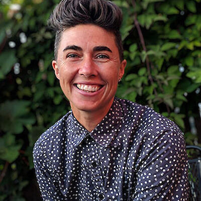Katrina Spade, smiling, with short dark hair styled up, wearing a polka-dot shirt, in front of green foliage