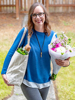 Laura Malcolm, smiling, wearing glasses and a blue sweater, holding a canvas grocery tote and a bouquet of flowers outdoors
