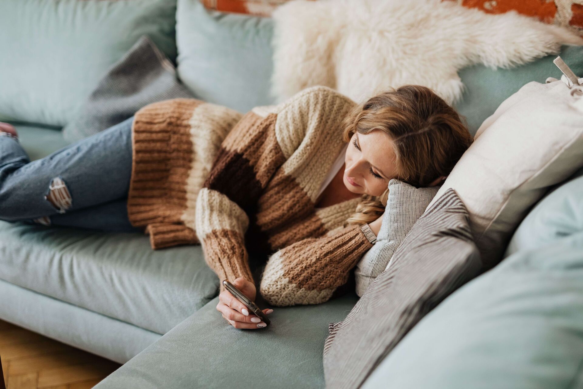 A woman resting comfortably on a couch in a cozy sweater, quietly reading a supportive text message on her phone, representing accessible, low-energy grief support at home.