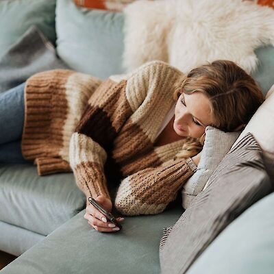 A woman resting comfortably on a couch in a cozy sweater, quietly reading a supportive text message on her phone, representing accessible, low-energy grief support at home.