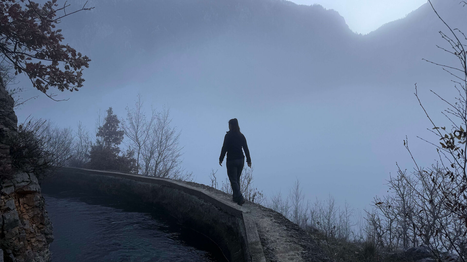 A silhouette of a person walking along a narrow concrete ledge next to water, surrounded by heavy mountain fog, symbolizing the complex and unresolved path of grieving a complicated relationship.