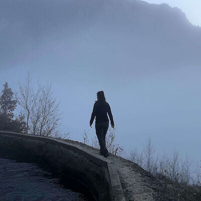A silhouette of a person walking along a narrow concrete ledge next to water, surrounded by heavy mountain fog, symbolizing the complex and unresolved path of grieving a complicated relationship.