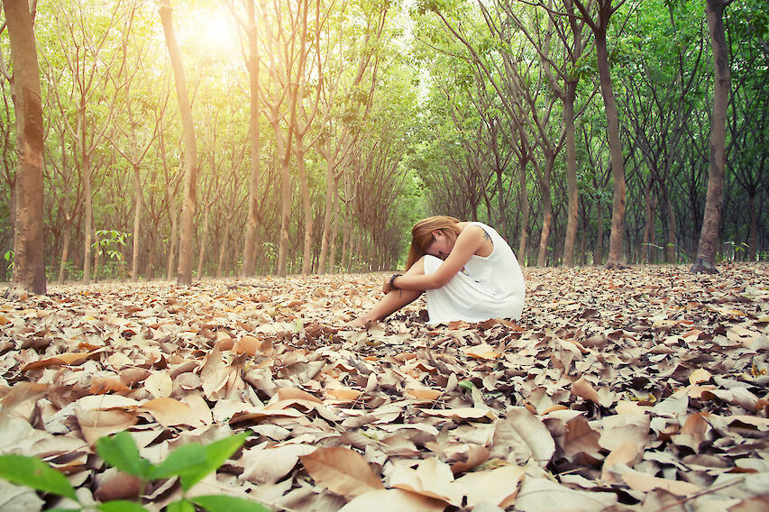 A woman in a white dress sitting alone with her head bowed on her knees, among fallen autumn leaves in a wooded grove