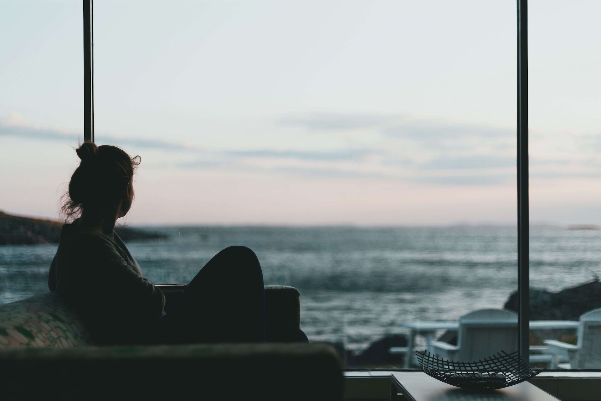 A silhouette of a person sitting quietly by a large window looking out at the ocean, representing the quiet reflection needed when navigating what grief is and coping with loss.