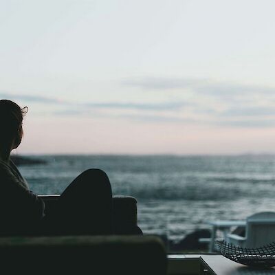 A silhouette of a person sitting quietly by a large window looking out at the ocean, representing the quiet reflection needed when navigating what grief is and coping with loss.