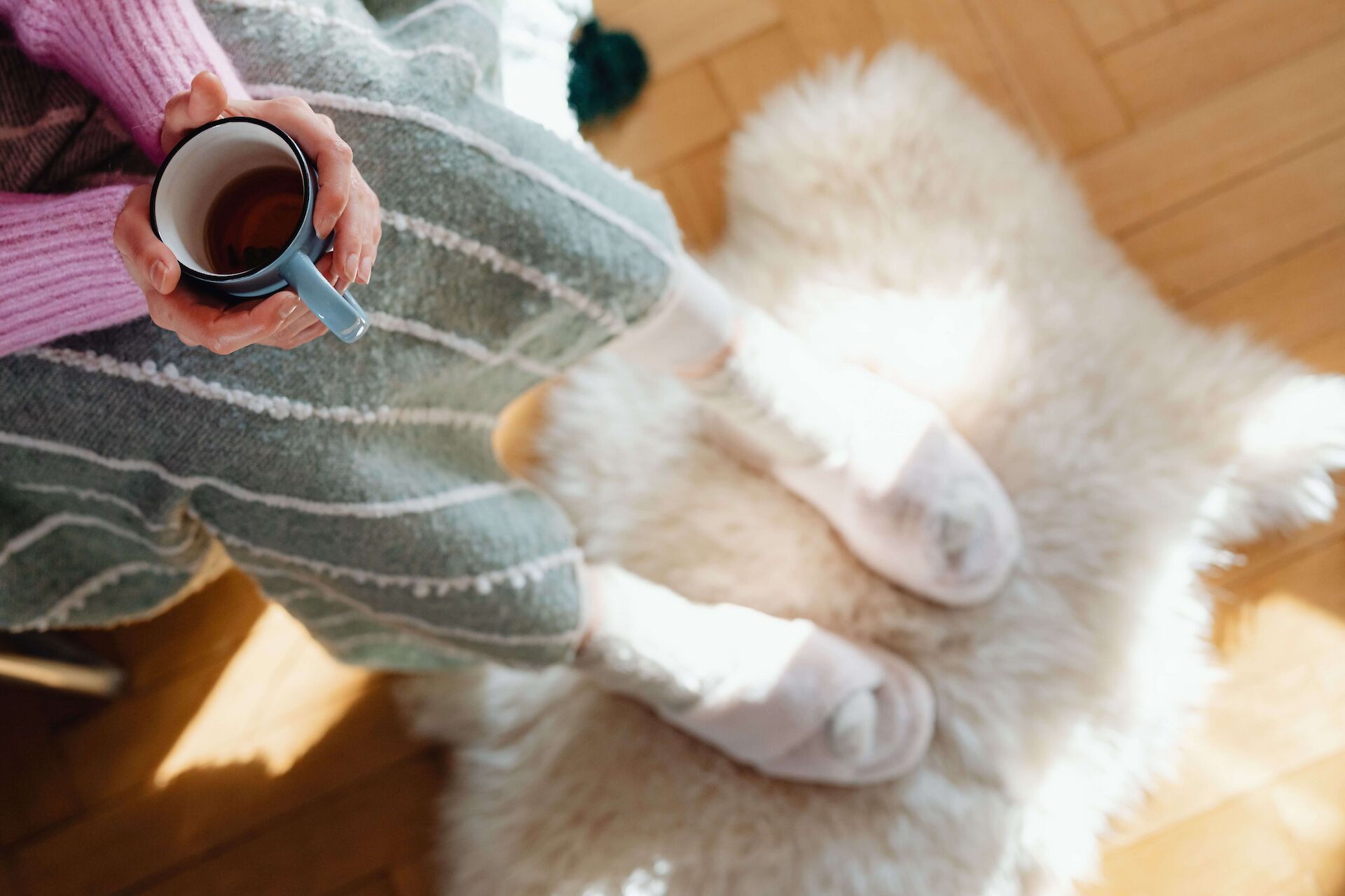 A top-down view of a person wearing cozy clothes and slippers, resting their feet on a fluffy rug in the sunlight while holding a warm mug. This represents a much-needed moment of quiet rest and self-care for a caregiver grieving a cancer loss.