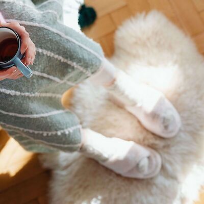 A top-down view of a person wearing cozy clothes and slippers, resting their feet on a fluffy rug in the sunlight while holding a warm mug. This represents a much-needed moment of quiet rest and self-care for a caregiver grieving a cancer loss.
