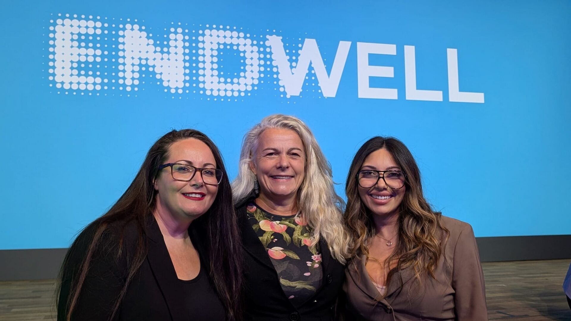 Three women smiling together in front of an Endwell conference screen