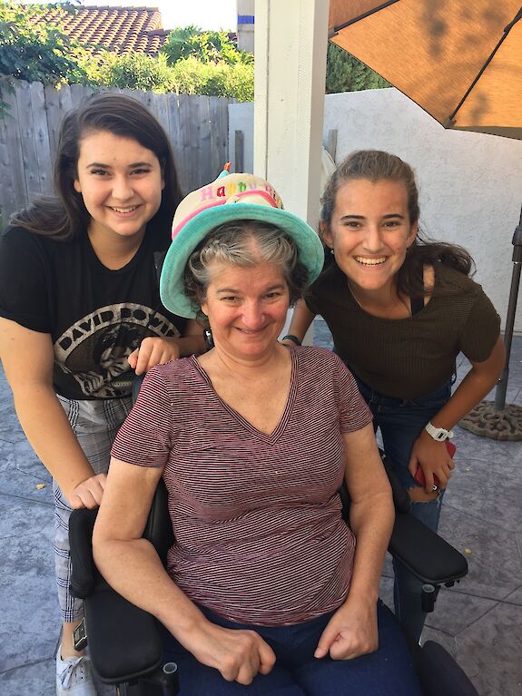 Renee's mother, wearing a Happy Birthday party hat and smiling, seated between her two teenage daughters outdoors