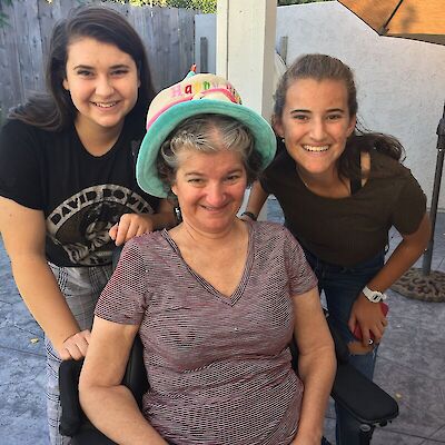 Renee's mother, wearing a Happy Birthday party hat and smiling, seated between her two teenage daughters outdoors