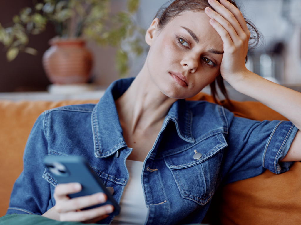 A young woman in a denim jacket sits on a sofa looking distressed, holding her phone with a hand to her forehead