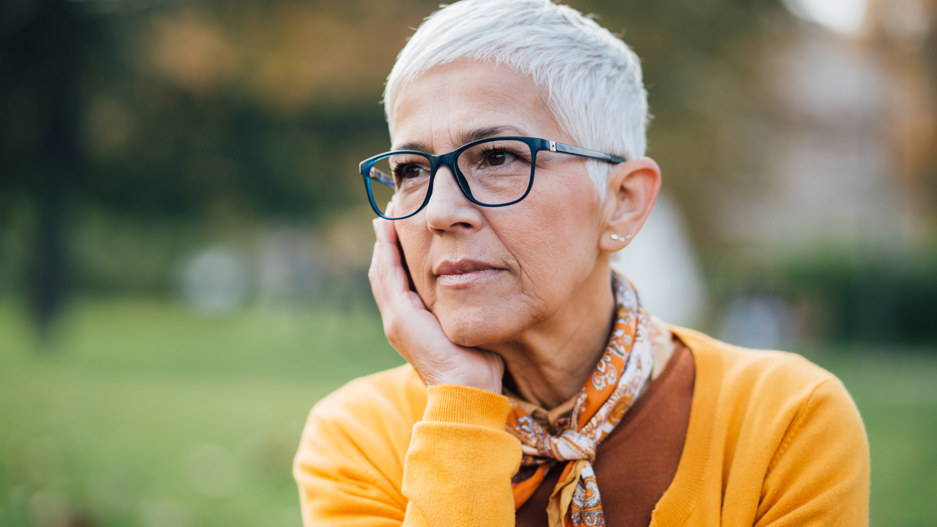 An older woman with glasses and an orange cardigan sits outdoors looking pensive