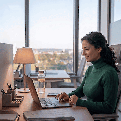A woman works at a laptop at a desk by a window with a city view