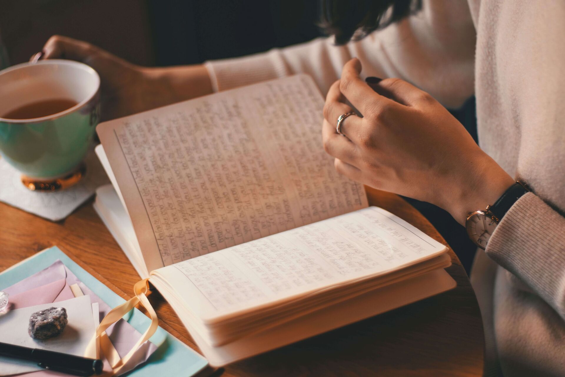 A close-up of a person’s hands resting on an open handwritten journal next to a warm cup of tea on a wooden table, taking time for quiet reflection during the grieving process.