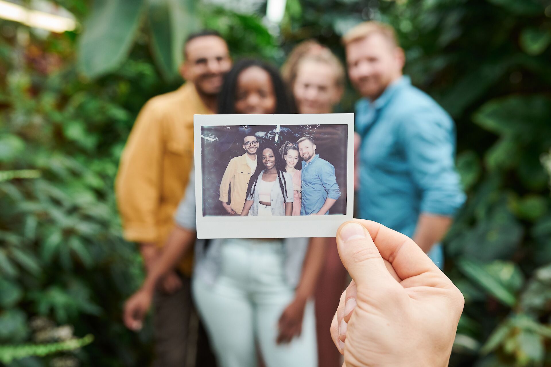 A hand holding up a polaroid photograph of four smiling friends with a blurred background of a greenhouse, representing the cherished memories that remain during grief after losing a friend.
