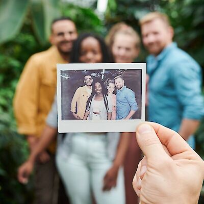 A hand holding up a polaroid photograph of four smiling friends with a blurred background of a greenhouse, representing the cherished memories that remain during grief after losing a friend.