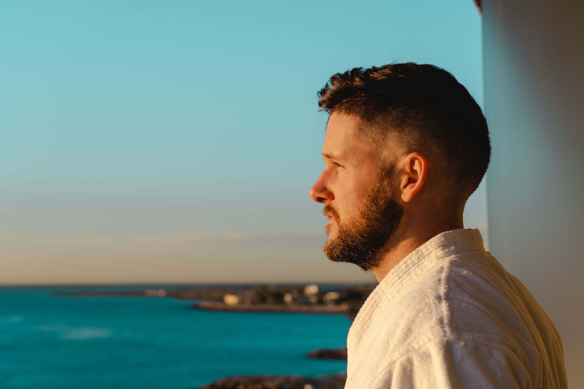 A side profile of a thoughtful adult man in a white robe looking out over the ocean at sunset, reflecting on memories and navigating the grief of losing your dad.