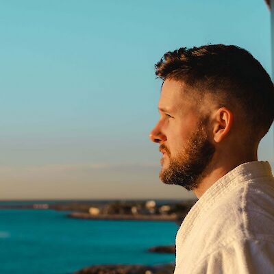 A side profile of a thoughtful adult man in a white robe looking out over the ocean at sunset, reflecting on memories and navigating the grief of losing your dad.