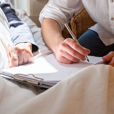 A caregiver sits beside a person resting in bed, writing notes on a clipboard
