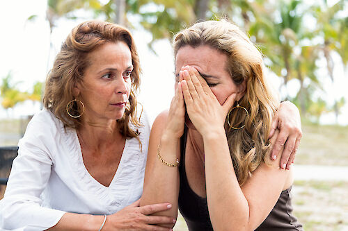 One woman comforting another who is crying with her hands over her face, seated outdoors
