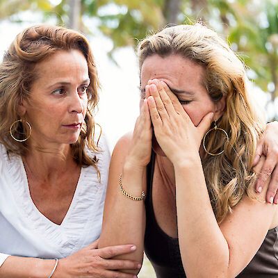 One woman comforting another who is crying with her hands over her face, seated outdoors